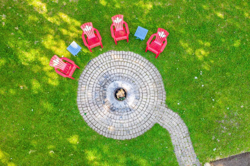 Aerial view of a circular fire pit area with red Adirondack chairs and small tables on a green lawn at Belwood Lake.